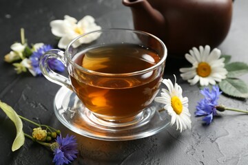 Aromatic herbal tea in glass cup, different flowers and teapot on black table, closeup