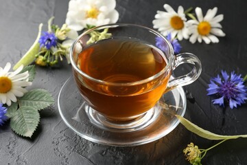 Aromatic herbal tea in glass cup, different flowers and green leaves on black table, closeup