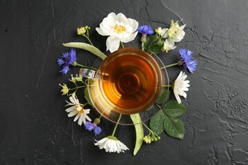 Aromatic herbal tea in glass cup, different flowers and green leaves on black table, flat lay