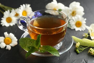 Aromatic herbal tea in glass cup, different flowers and mint on black table, closeup