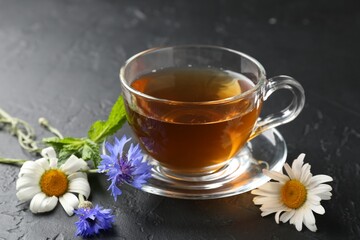 Aromatic herbal tea in glass cup, different flowers and mint on black table, closeup