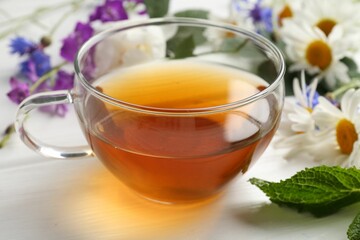 Aromatic herbal tea in glass cup, different flowers and mint on white wooden table, closeup