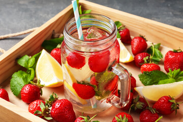 Refreshing drink with strawberries, lemon and mint in mason jar surrounded by fruits in wooden crate on grey table, closeup