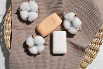 Soap bars, towel and cotton flowers in wicker basket on white table, top view