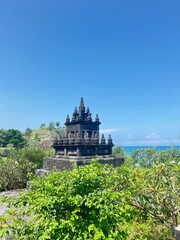 Coastal Temple Shrine: A serene coastal temple, perched atop a verdant hill, overlooks a tranquil ocean under a clear blue sky. The ancient stone structure, detailed with intricate carvings.