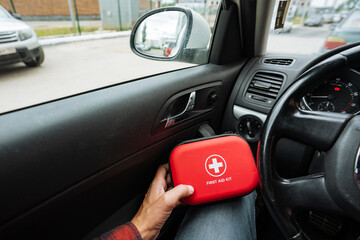 A person is comfortably seated in a car, firmly holding onto a bright red first aid kit, which is...