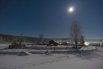 Snow-covered landscape at night, illuminated by a bright moon, featuring a rustic wooden house and...