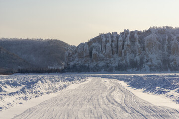 Snow-covered road winding through a winter landscape, flanked by towering rock formations and dense...