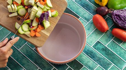 A female hand gently pours freshly chopped vegetables — including zucchini, carrot, pepper, and...