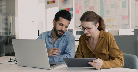 Diverse couple of company colleagues using laptop and tablet together. Friendly advisor, mentor showing work application, software on digital gadget to new employee, giving help to coworker - Powered by Adobe