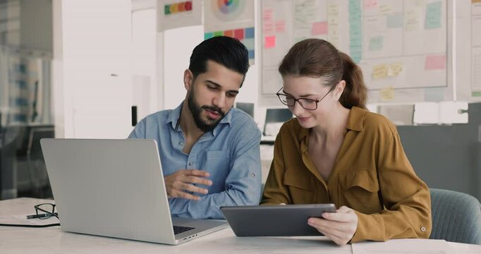 Diverse couple of company colleagues using laptop and tablet together. Friendly advisor, mentor showing work application, software on digital gadget to new employee, giving help to coworker