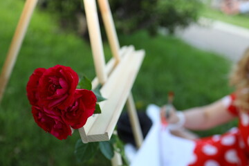 A bright red rose sits on an easel as an artist sketches its beauty. The scene takes place in a...