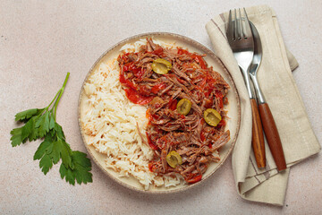 Ropa vieja, a traditional Cuban dish, is served with white rice, tomato sauce, and green olives on a plate from above on light beige background