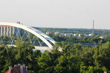 White arch bridge spans over a river, surrounded by lush greenery and industrial buildings in the background on a clear day.
