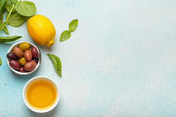 Lemon, olive oil, olives and fresh basil leaves arranged on a light blue textured background create...