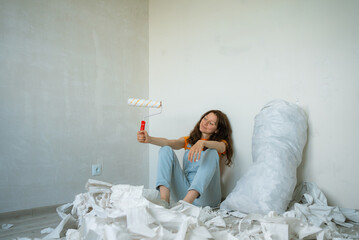 Young woman taking a break during apartment renovation, holding a paint roller and sitting on the...