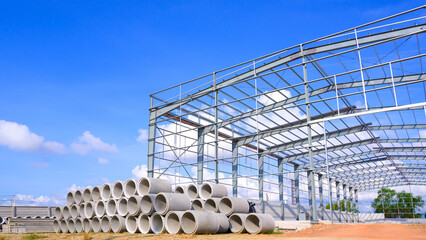 Metal structure of warehouse industrial factory building with concrete drainage pipes stacked on the ground in construction site against blue sky background, perspective side view 