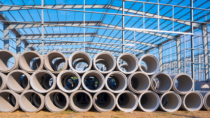 Concrete drainage pipes stacked on the ground in front of metal structure of warehouse industrial factory building in construction site, front view 