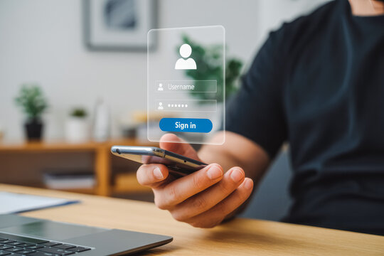 A person uses a smartphone to access a login interface, featuring username and password fields. A laptop and blurred plant in the background suggest a modern workspace environment.