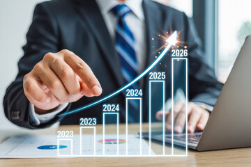 Businessman in a suit points to a rising graph with bars labeled 2023 to 2026, symbolizing growth. The scene conveys strategic business planning and success projection on a laptop.