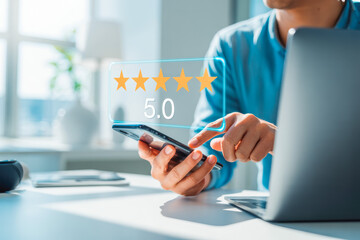 A person uses a smartphone to rate a service with five stars, displaying a 5.0 rating review feedback. The setting includes a laptop, signifying digital engagement and satisfaction.