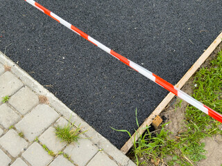 Fresh asphalt paving is being installed beside an existing sidewalk in a public space. Caution tape...