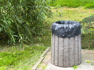 A concrete trash bin with a black liner stands in a grassy area amidst greenery, catching sunlight...