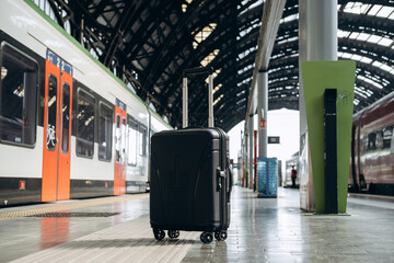 Black hard-shell suitcase positioned on platform at modern train station, with trains in background...