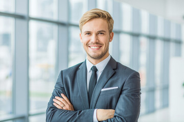 Professional businessman in a smart suit stands confidently in a modern office. He smiles warmly, with arms crossed, exuding success and authority. Bright background emphasizes his charisma.