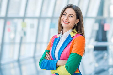 A confident woman in a vibrant, colorful jacket smiles warmly, standing in a modern, bright office space. Her attire reflects creativity and positivity, enhancing the vibrant workplace atmosphere.