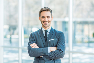 Professional businessman in a tailored suit stands confidently with arms crossed in a bright office setting. Smiling, with natural light pouring in, enhancing a crisp, modern ambiance.