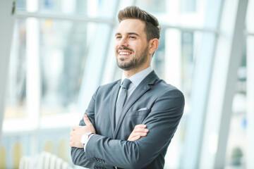Confident businessman in suit with folded arms, smiling in a modern office space, exuding professionalism and success. Bright natural light and sleek interior design enhance the ambiance.