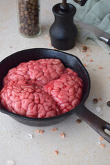 Whole beef brain in black pan on kitchen table beside salt and pepper grinder. Rustic culinary concept with focus on raw texture and overhead preparation of organ meat.