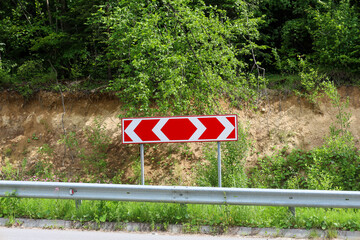 A bright road sign indicating a sharp turn is located by the roadside. Behind it, green trees and...
