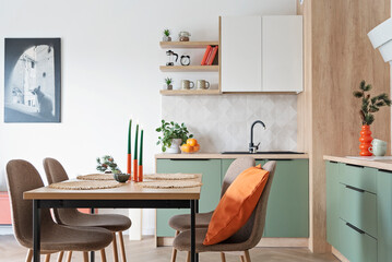 Modern kitchen and wooden dining table with chairs. Green furniture, beige tiles on the wall and orange decoration. New home with elegant design and picture on the wall. Real estate.