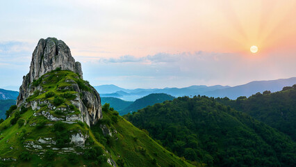 Scenery of a beautiful sunset with a dominant rocky mountain covered in greenery. A distant view of the mountains in the sunlight of the sun and rays. Background, wallpaper.
