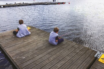 Two boys fishing while sitting on wooden pier at calm lake on sunny summer day. Sweden.