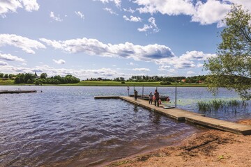 Family walking on wooden pier by calm lake with moored boats on summer day. Sweden.