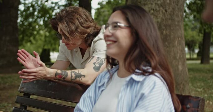 Young carefree friends are enjoying a relaxed conversation on a park bench outdoors. They share smiles and laughter, embodying a happy and social connection in a vibrant green setting during summer.