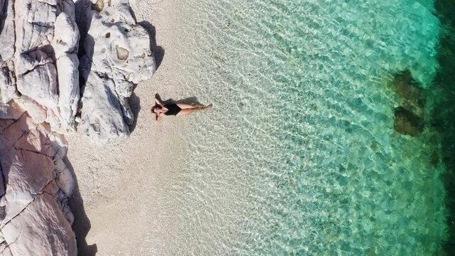4K drone footage of woman dressed black swimsuit prostrated on white pebble beach lying down on the calm Ionian sea waves. Exotic tropical summer holidays concept. Cephalonia island, Greece.