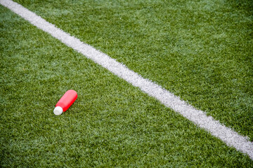 red water bottle lies on the green artificial grass of a football field near the dividing white line, the concept of drinking during long workouts and replenishing fluids and electrolytes,