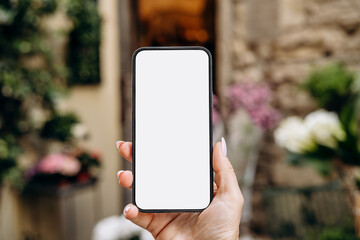 Mockup woman's hand holding using smart phone with white blank screen POV surrounded by soft floral...