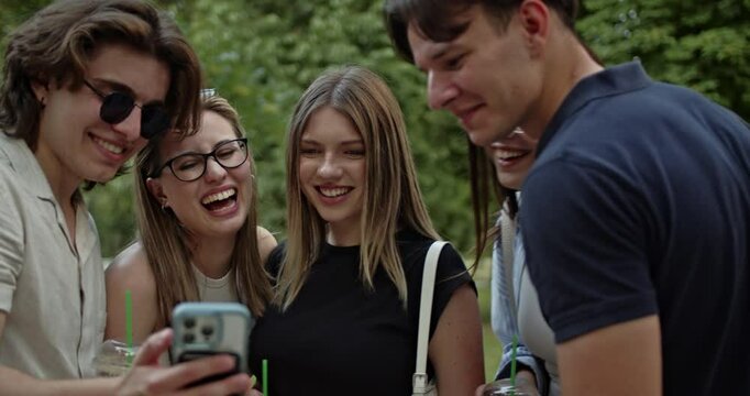 A diverse group of carefree friends shares a moment of joy and laughter, gathered closely around a smart phone outdoors. They are enjoying each other's company while watching content on the device.