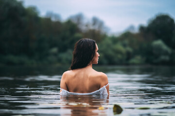 Fototapeta premium Beautiful long-haired brunette in the lake in a white dress. Portrait of a woman back view in the water in summer cloudy weather