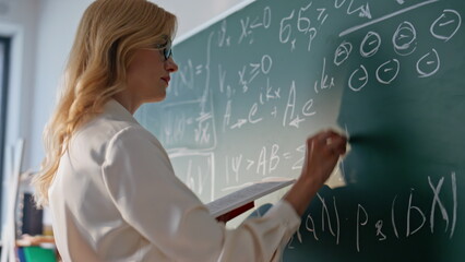 Teacher writing math equations on chalkboard looking book closeup. Woman tutor 