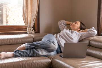 A woman stretches her neck while lying on the couch in front of her laptop