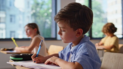 Elementary schoolboy writing notes sitting class closeup. Cute boy laughing