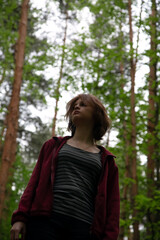 Naklejka premium Beauty face young woman looking up on a green pine trees in the forest on sun spring weather. Girl walking in woods, eyes look above in fashion clothes and breath fresh air. Closeup emotion portrait