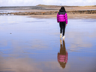 Teenager girl in purple jacket walking on a wet sandy beach which reflects blue cloudy sky. Travel to seaside concept. Relaxing activity by the ocean. Selective focus.