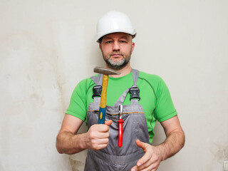 A workman in a green shirt and gray overalls is holding tools Portrait of a male general laborer on a construction site in white safety helmet. Model in his 40s with grey beard.